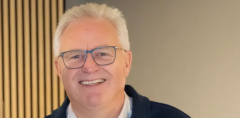 Person mit weißem Haar und Brille vor goldener Lamellenwand, trägt dunkle Jacke und helles Hemd, frontal aufgenommen.